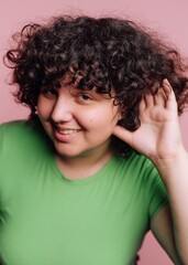 Young woman with curly hair playfully poses with hand to ear against pink backdrop