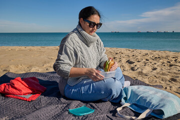 Woman enjoying a homemade sandwich while sitting on a blanket at a beach picnic.