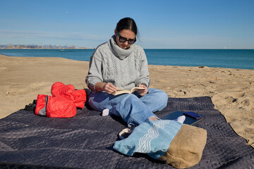 Woman relaxing on a beach blanket while reading a book on a sunny day by the sea.