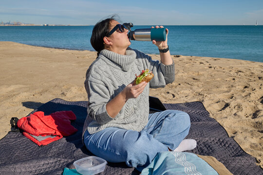 Woman drinking from a thermos while eating a sandwich at a beach picnic on a sunny day. - Powered by Adobe