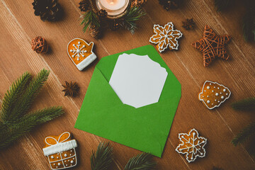 Green envelope with blank card surrounded by festive Christmas cookies and pine on wood