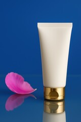 White cosmetic tube standing upright on white surface with pink rose petals and deep blue background, clean minimalist skincare packaging.
