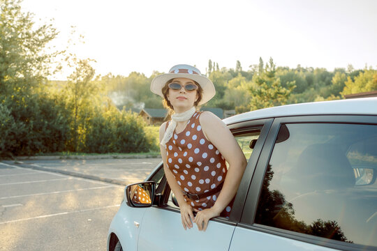 Joyful woman in vintage polka dot dress waving her straw hat from car window. Retro summer travel mood, freedom, happiness, and vacation adventure concept - Powered by Adobe