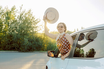 Joyful woman in vintage polka dot dress waving her straw hat from car window. Retro summer travel mood, freedom, happiness, and vacation adventure concept