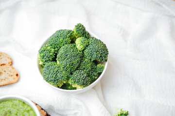 Fresh broccoli florets in white bowl with bread slices and green dip on white fabric