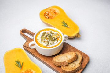 Orange soup in bowl with seeds and bread on wooden board, butternut squash halves on side