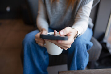 Closeup of a person sitting in a cozy cafe holding a smartphone and a white coffee mug, relaxed casual atmosphere with soft natural light and warm lifestyle vibes.