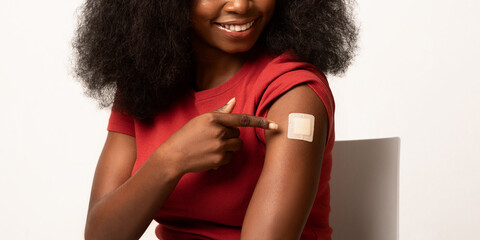 A happy African American woman sits against a white background, pointing at the sticking plaster on her arm after receiving a Covid-19 vaccine. She promotes vaccination and health awareness.