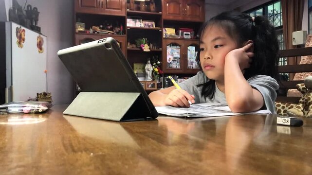 Asian young girl sits at a wooden table, bored, engaged in online study with a tablet and workbook, pencil in hand. The room is cozy, with cabinets and various decorations in the background
