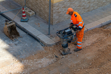 Construction worker in orange safety gear operating a tamping rammer machine to compact gravel in a trench. Road maintenance scene with an excavator bucket and traffic cone nearby.