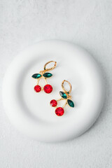 Elegant cherry earrings with red gemstones and green leaves placed on white round plate on light background