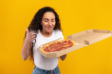 A joyful Latin lady stands with a pizza delivery box, savoring the aroma of tasty Italian pizza. She has closed eyes, expressing excitement for her delicious meal on a bright yellow orange background.