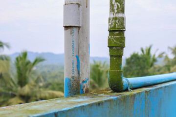 Algae-covered PVC water pipes on a rooftop tank with hill and coconut tree background, showing weathered plumbing and outdoor exposure.