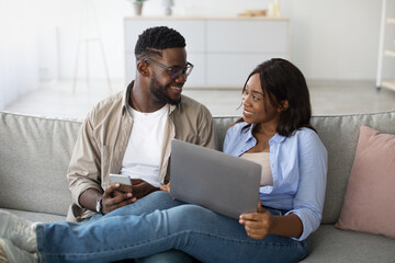 Happy black couple sits closely on their couch, interacting with a smartphone and laptop. They are relaxing at home, sharing laughter and enjoying each other’s company while browsing online.