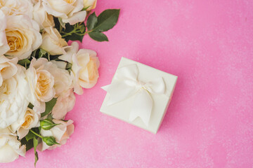 White gift box with ribbon surrounded by roses on pink background