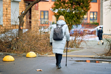Woman with cane walking through wet residential courtyard where melting snow, puddles, and winter...