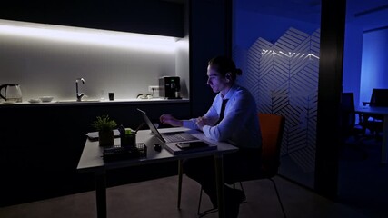 A focused businessman is diligently working on his laptop late at night in a quiet, dimly lit office kitchen. - Powered by Adobe