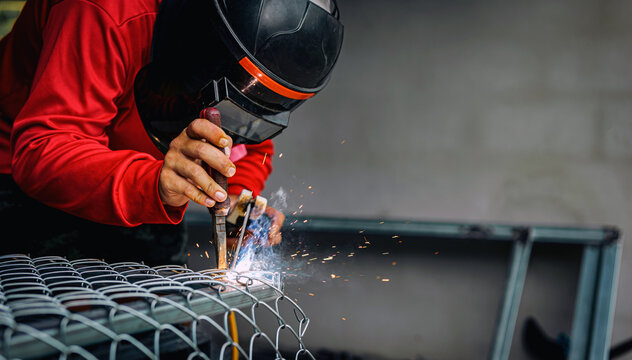 Welder wearing a red long-sleeved shirt and a protective mask is welding a steel grating, Industrial Welder With Torch