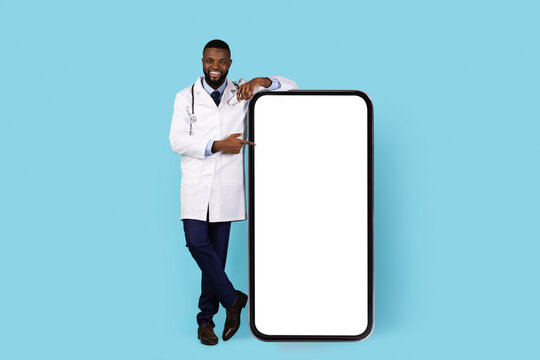 Smiling Black Doctor In Uniform Pointing At Big Blank Phone With White Screen, African American Therapist Demonstrating Copy Space For Online Medical App Design, Blue Studio Background, Mockup - Powered by Adobe