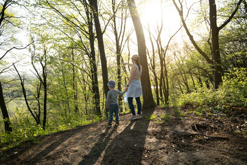 Obraz premium Young mother and four year old toddler son looking at beautiful view standing in forest. Spending time together in beautiful warm spring evening. Back view. Lovely moment.