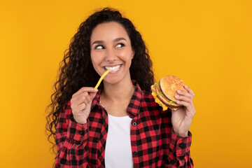 A joyful woman bites into a delicious burger, holding a fry in her other hand. She looks excited,...