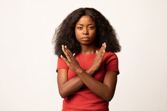 A serious millennial African American woman stands displaying a stop gesture with her arms crossed. She is posing against a plain white studio background, conveying a clear message of refusal.