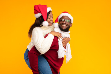 A joyful African American couple celebrates Christmas by having fun together. The husband carries his wife piggyback while they wear Santa hats, posing cheerfully against a yellow background.