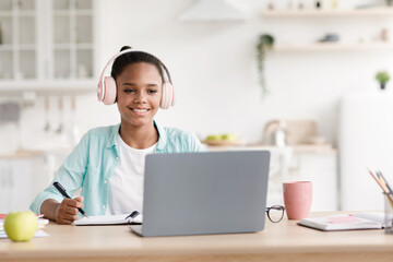 A happy African American schoolgirl listens to a virtual lesson using her laptop at home. She takes notes in her kitchen while wearing earphones, enjoying the learning experience.