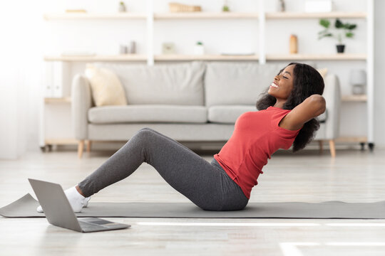 A woman is doing a fitness workout at home, using a laptop for guidance. She is smiling while performing an exercise on a mat in a well-lit living room.
