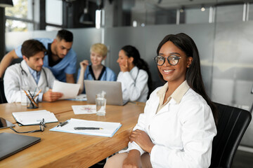 Fototapeta premium Pretty african american young lady doctor smiling at camera while attending breefing, international team of doctors sitting at table at clinic, looking at laptop screen, discussing medical cases