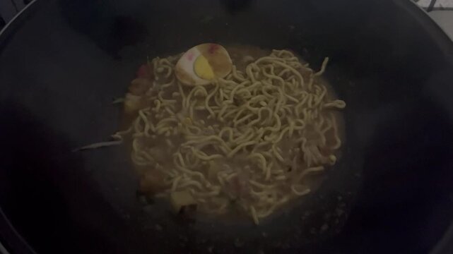 Boiled noodles are being heated in a hot pan. The texture of the noodles is soft and cooked, creating a warm and appetizing cooking atmosphere.