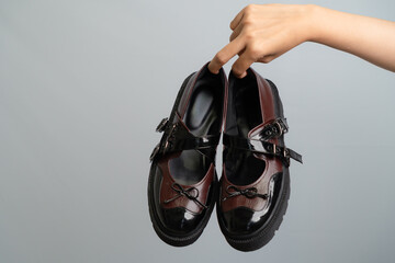 Hand Holding A Pair Of Brown And Black Mary Jane Shoes Against A Gray Background With Soft Lighting