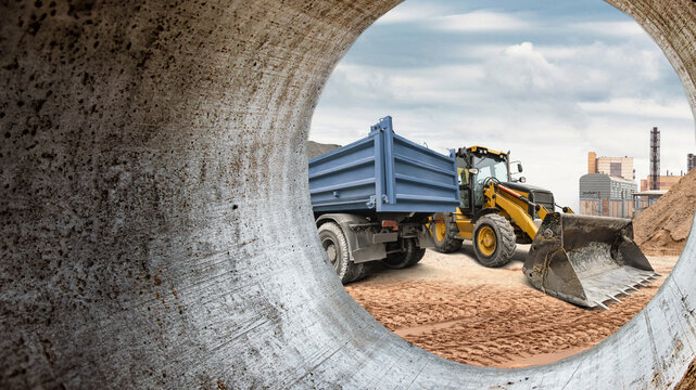 Heavy machinery operates at a construction site in an urban area. A dump truck and a loader work on moving materials under a cloudy sky