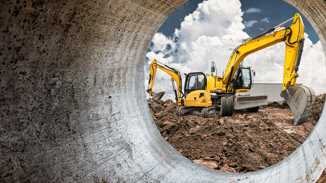 Two yellow excavators are digging and maneuvering dirt on a construction site viewed through a large concrete pipe under a blue sky with clouds