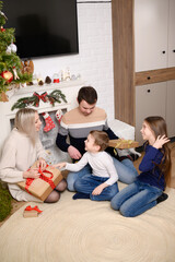 Happy family sits on floor near fireplace. Mom gives Christmas gifts to children and husband at home on Christmas Eve near decorated tree. Concept of celebrating Christmas and New Year, gifts