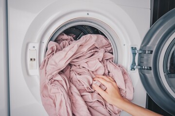A Hand Inserting Soft, Pink Bedding Into a Modern Washing Machine or Clothes Dryer for Fresh...