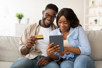 Millennial African American couple relaxes on their couch, excitedly exploring online shopping. The guy holds a debit card while they choose items on a tablet, sharing joyful moments together.