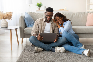 Obraz premium In a warm and inviting living room, a young black couple sits on the floor, engaged with a laptop.