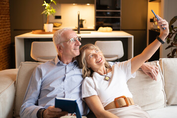 Happy senior couple relaxing on sofa, taking a selfie with smartphone in modern home