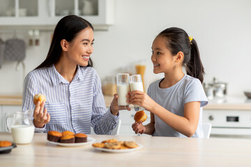 In a bright kitchen, a girl and her mother share a joyful moment clinking glasses filled with milk. They smile at each other while enjoying cookies and cakes, creating a warm atmosphere.