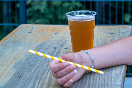 Hand holding a striped paper straw next to a plastic cup of beer outdoors on wooden table - Powered by Adobe