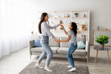 In a bright living room, an asian mom and her daughter are happily dancing while holding hands. They are enjoying their favorite song together, creating joyful memories at home.