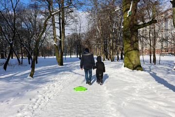 Man walking with child and pulling green sled in snowy urban park. Winter outdoor activity.