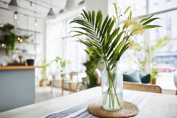 Close-up, a glass vase with palm leaves and white flowers stands on a table with a woven placemat. A bright, modern cafe interior, in the background. Cozy interior design. Cafe bistro.