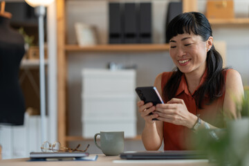 Focused Asian woman entrepreneur using her smartphone while working at a laptop in a cozy home...