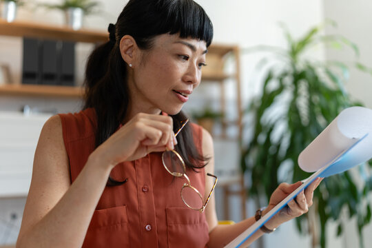 Asian woman small business owner reviewing documents in her home office, holding her glasses and analyzing paperwork with a thoughtful expression