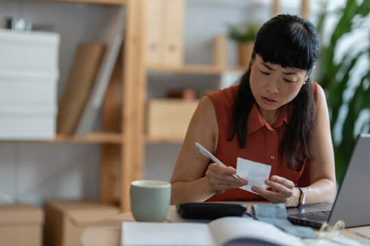 Asian woman entrepreneur in her home office, checking receipts and entering numbers on her phone while working with a laptop and calculator at a round desk