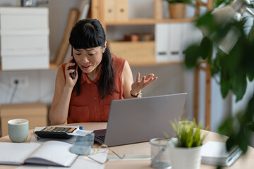 Fototapeta premium Asian woman small entrepreneur stressed while handling finances in her home office, talking on the phone and checking bills with a laptop and calculator on the desk