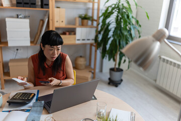 Asian woman entrepreneur in her home office, checking receipts and entering numbers on her phone while working with a laptop and calculator at a round desk
