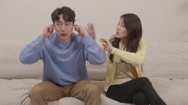 Asian couple arguing on the couch, woman nagging while man covers his ears
